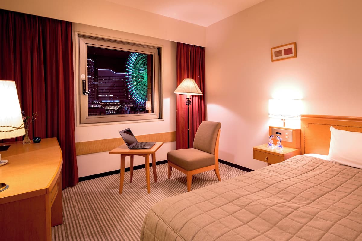 A hotel room with a bed, a desk with a laptop, a chair, a nightstand with a lamp, and a window with a view of a cityscape at night.
