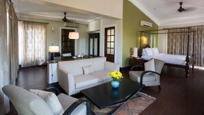 A hotel room with a bed, sofa, armchair, and a coffee table with flowers. There is a desk with a lamp and a mirror. - Sol De Goa by Sereno (Nerul)