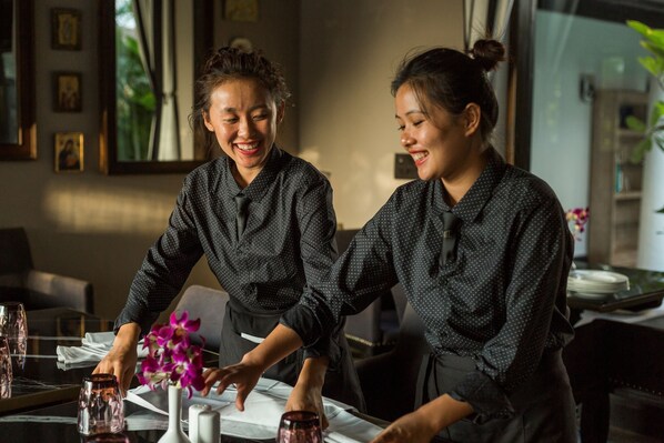 Two individuals in a restaurant setting, preparing a table with a white tablecloth and pink flowers.
