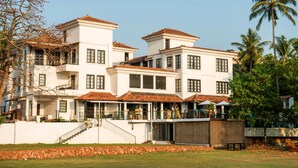 A large, multi-story building with a white facade and red-tiled roofs, featuring a covered outdoor seating area and a staircase leading to the entrance. - Sol De Goa by Sereno (Nerul)