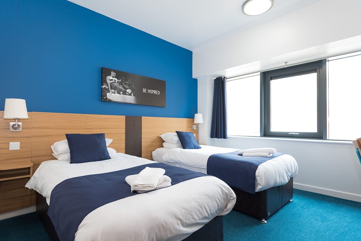 A hotel room with two beds, a blue accent wall, a framed inspirational quote, and a window with curtains.