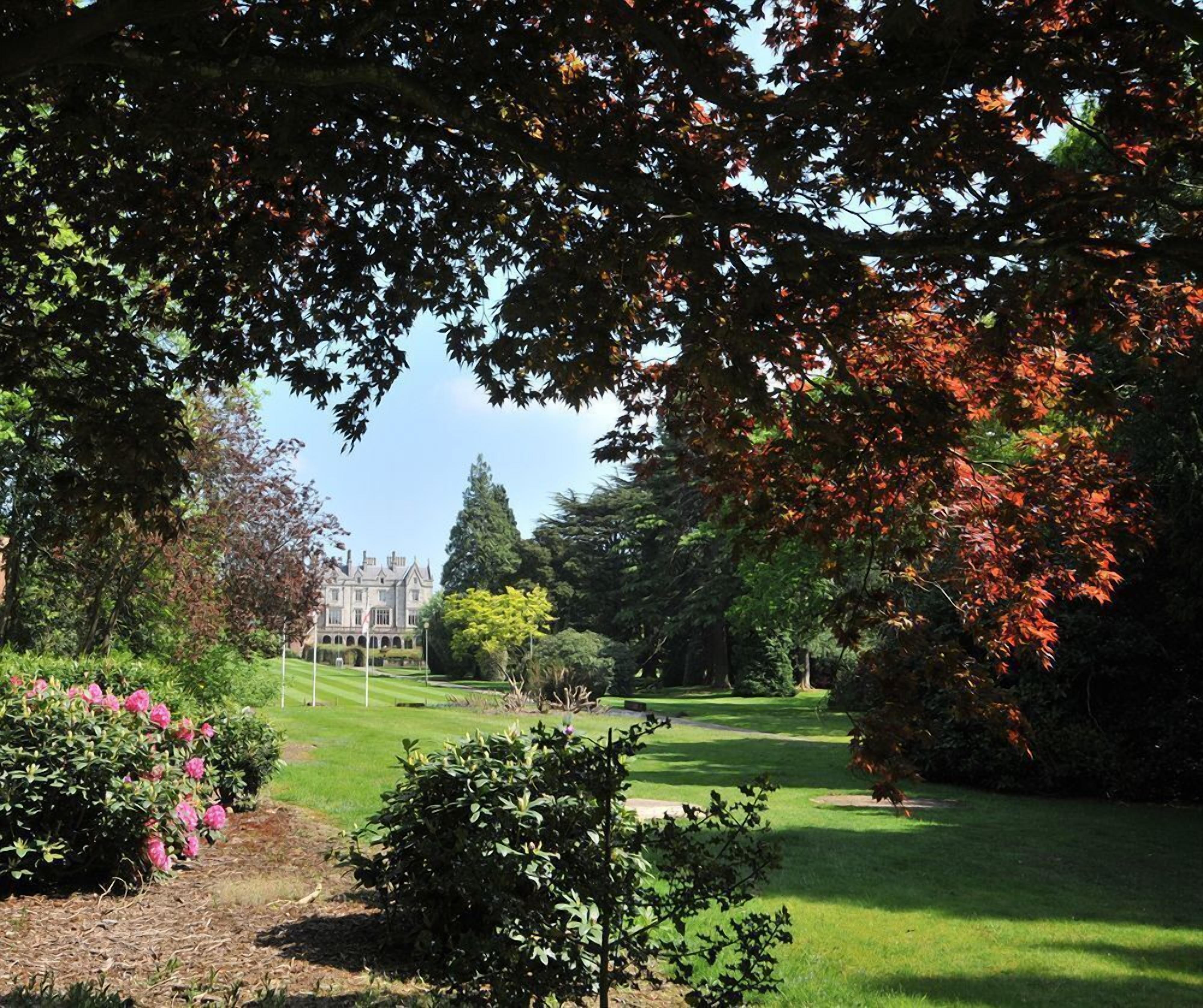 A well-maintained garden with a variety of trees, including some with autumn-colored leaves, a lawn, and a building in the background.