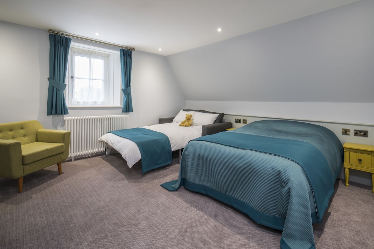 A bedroom with two beds, a window with curtains, a radiator, a yellow nightstand, and a green armchair.