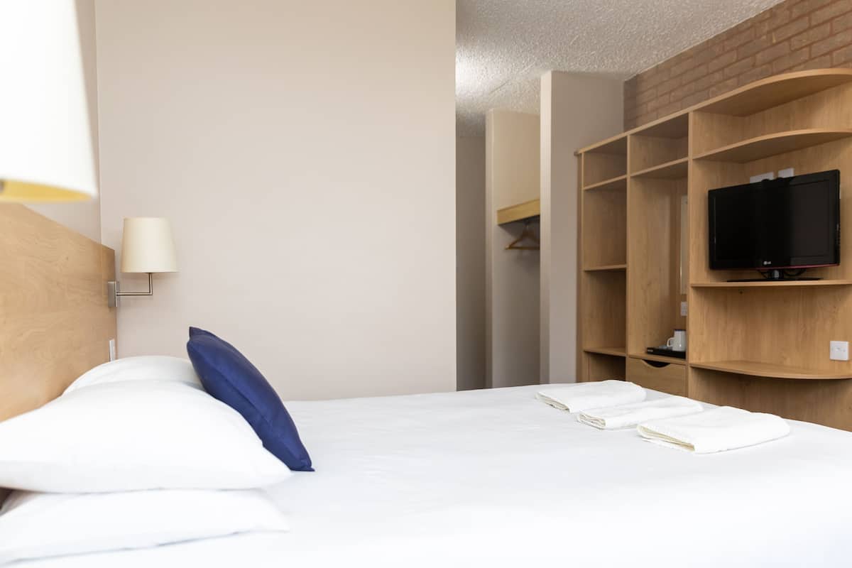 A hotel room with a bed, a TV on a wooden shelf, and a lamp on the wall.