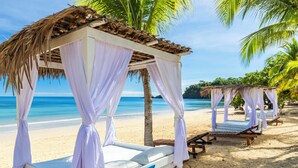 A beachside bed with a thatched roof and white curtains, a wooden bench, and a palm tree. - VOI Amarina Resort (Nosy Be)