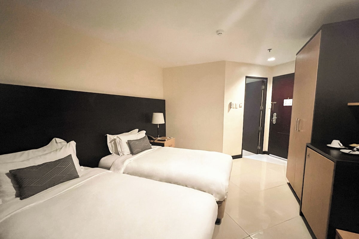 A hotel room with two beds, a nightstand with a lamp, a wardrobe, and a door leading to another room.