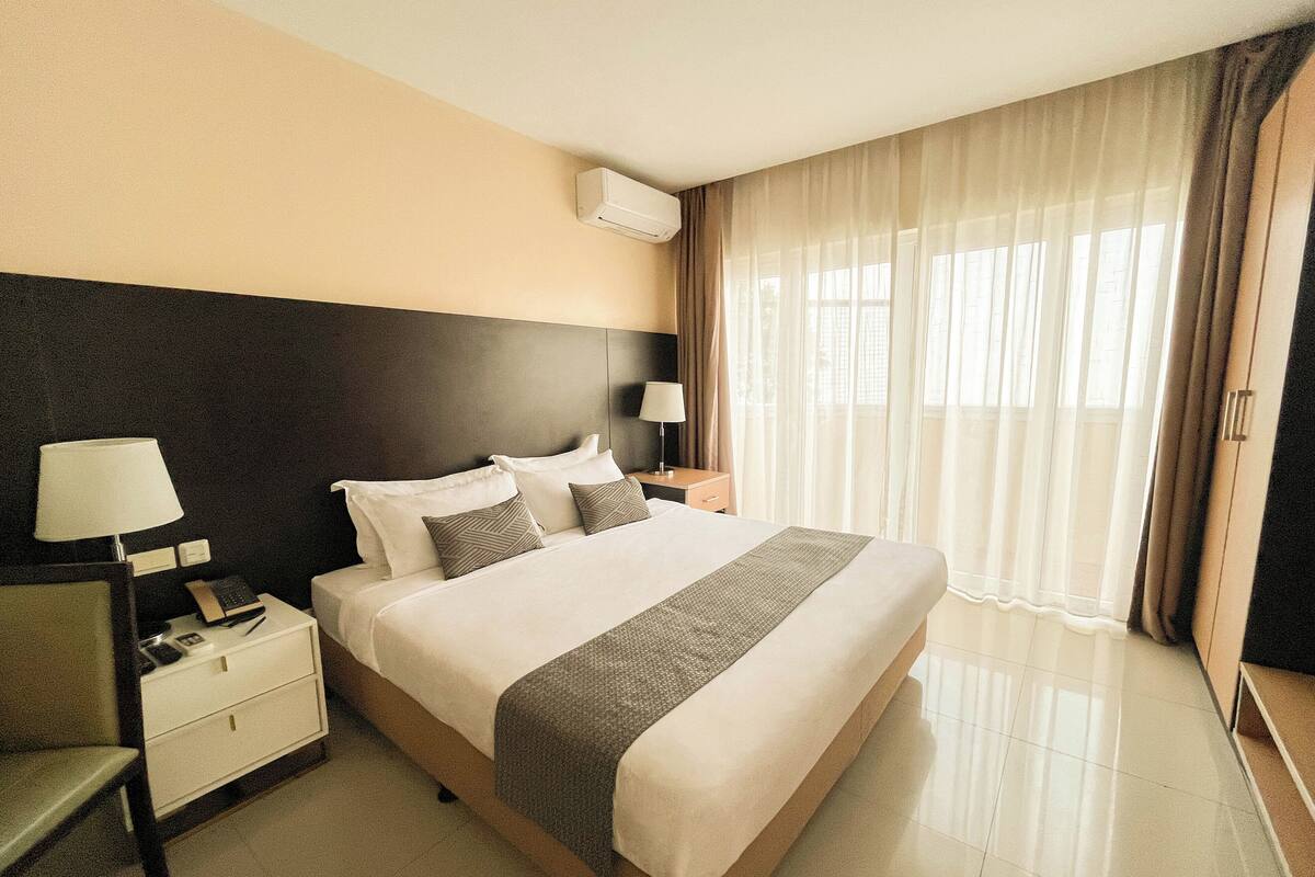 A hotel room with a large bed, a bedside table with a lamp, a nightstand, and a window with curtains.