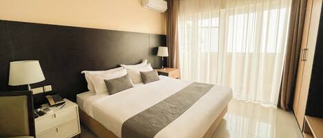 Une chambre d’hôtel avec un grand lit, une table de chevet avec une lampe, une commode et une fenêtre avec des rideaux.
