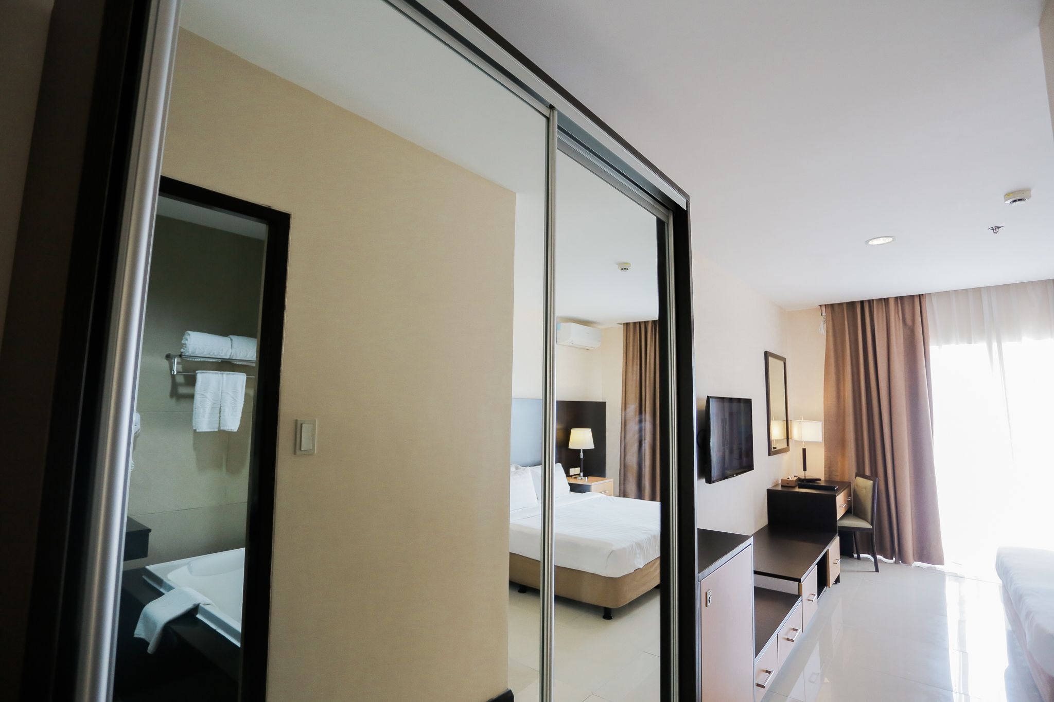 A hotel room with a bed, a desk, a chair, a TV, and a bathroom visible through a glass door.