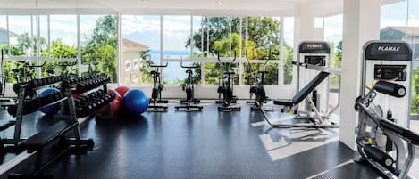 Un gimnasio con equipos de ejercicio variados, amplias ventanas y vistas a zonas verdes y un cuerpo de agua.