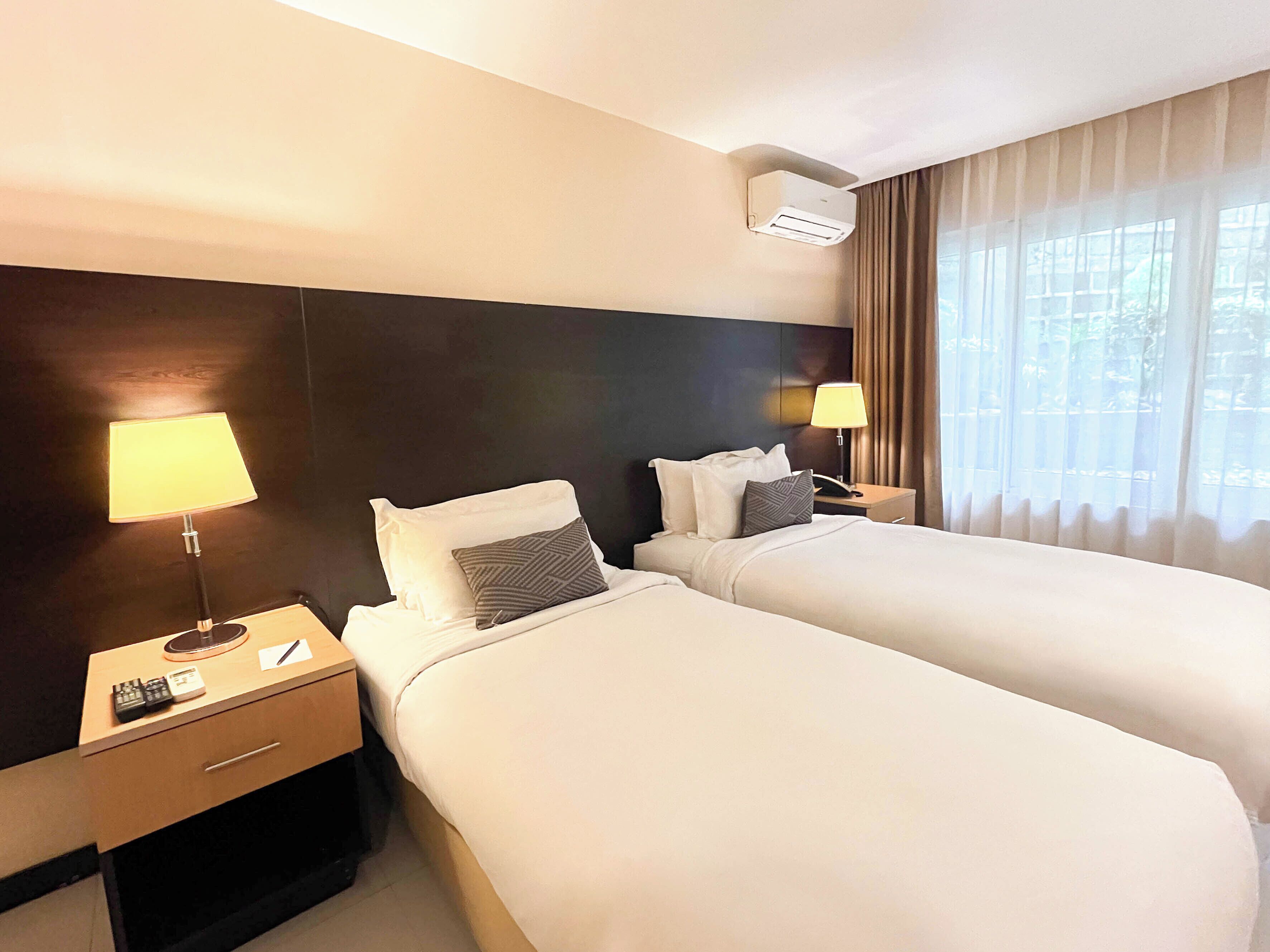 A hotel room with two beds, a nightstand with a lamp, a phone, and a remote control.