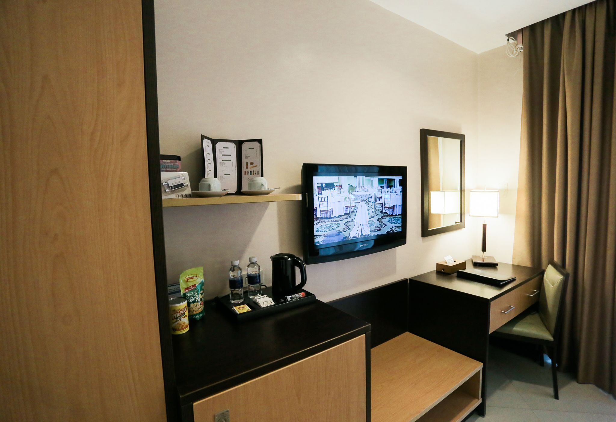 Habitación de hotel con televisión de pantalla plana montada en la pared, escritorio con lámpara, una silla y un estante pequeño con cafetera y aperitivos.