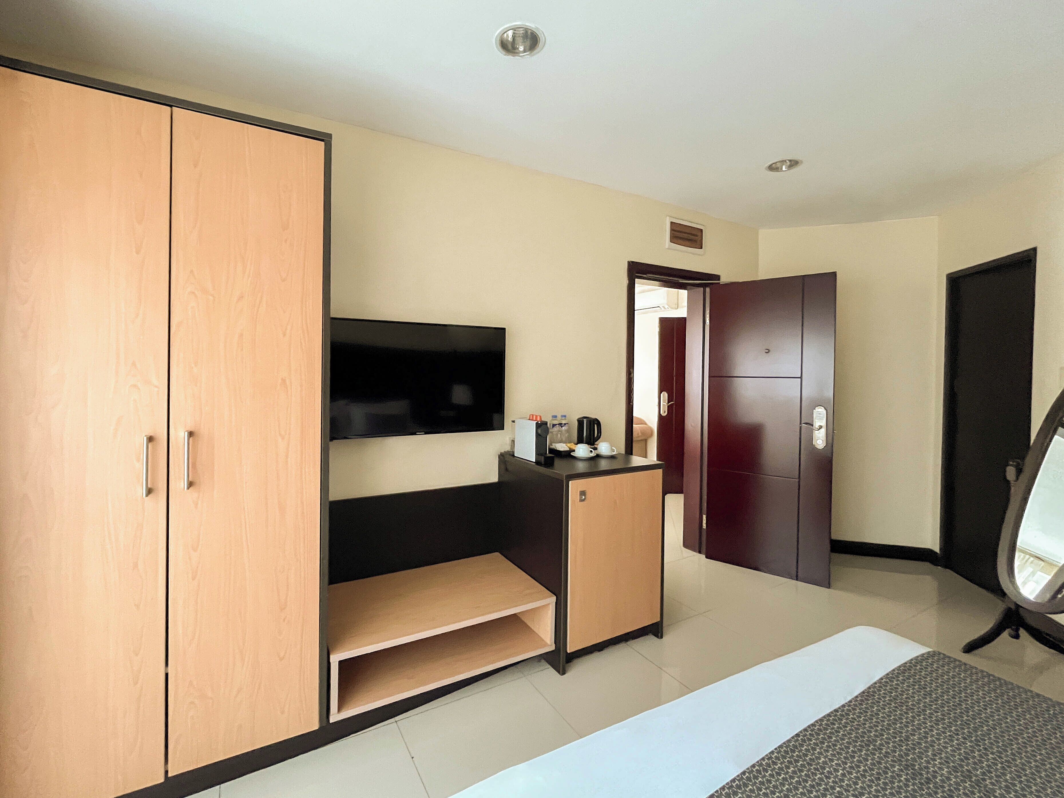 A hotel room with a large wardrobe, a small desk with a flat-screen TV, a coffee maker, and a door leading to another room.
