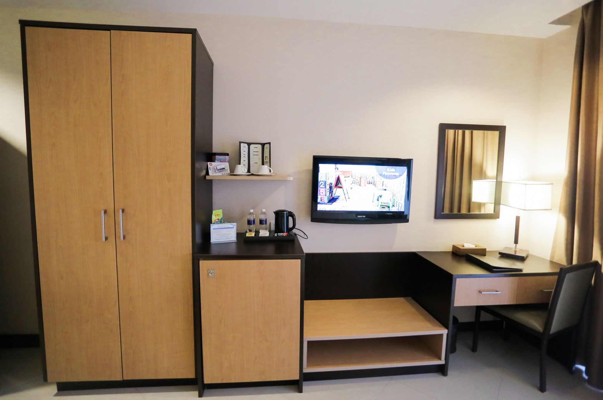 Une chambre d’hôtel avec une armoire en bois, un bureau avec une chaise, une télévision, un miroir et une lampe.