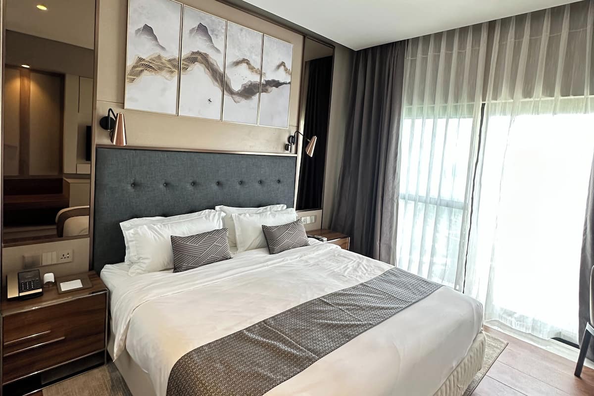 A modern hotel room with a large bed, a headboard with a decorative panel, a bedside table with a lamp, and a large window with curtains.