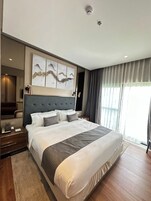 Egy modern szállodai szoba, amelyben egy nagy ágy található, a fejrészét díszes panel borítja, az ágy mellett egy éjjeliszekrény áll, rajta egy lámpával, és egy nagy ablak húzódik végig, melyet függönyök takarnak.