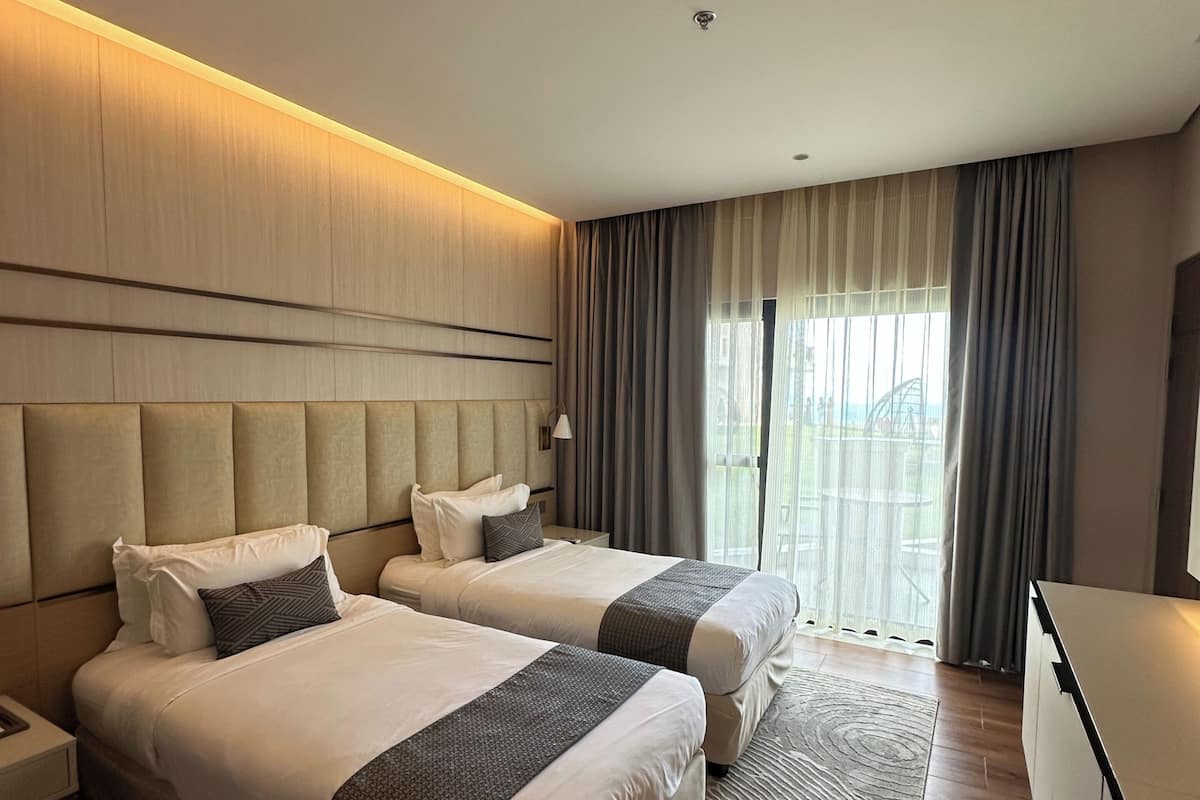 A hotel room with two beds, a desk, a chair, and a large window with curtains.