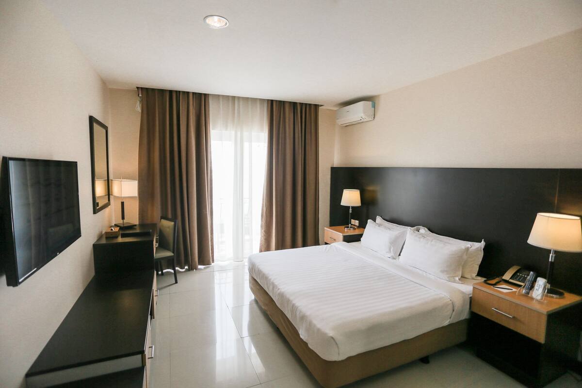 A hotel room with a large bed, bedside tables with lamps, a desk with a chair, a TV, and a window with curtains.