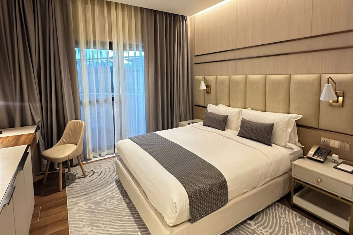 A hotel room with a large bed, bedside tables, a desk, a chair, and a view of the outdoors through the window.