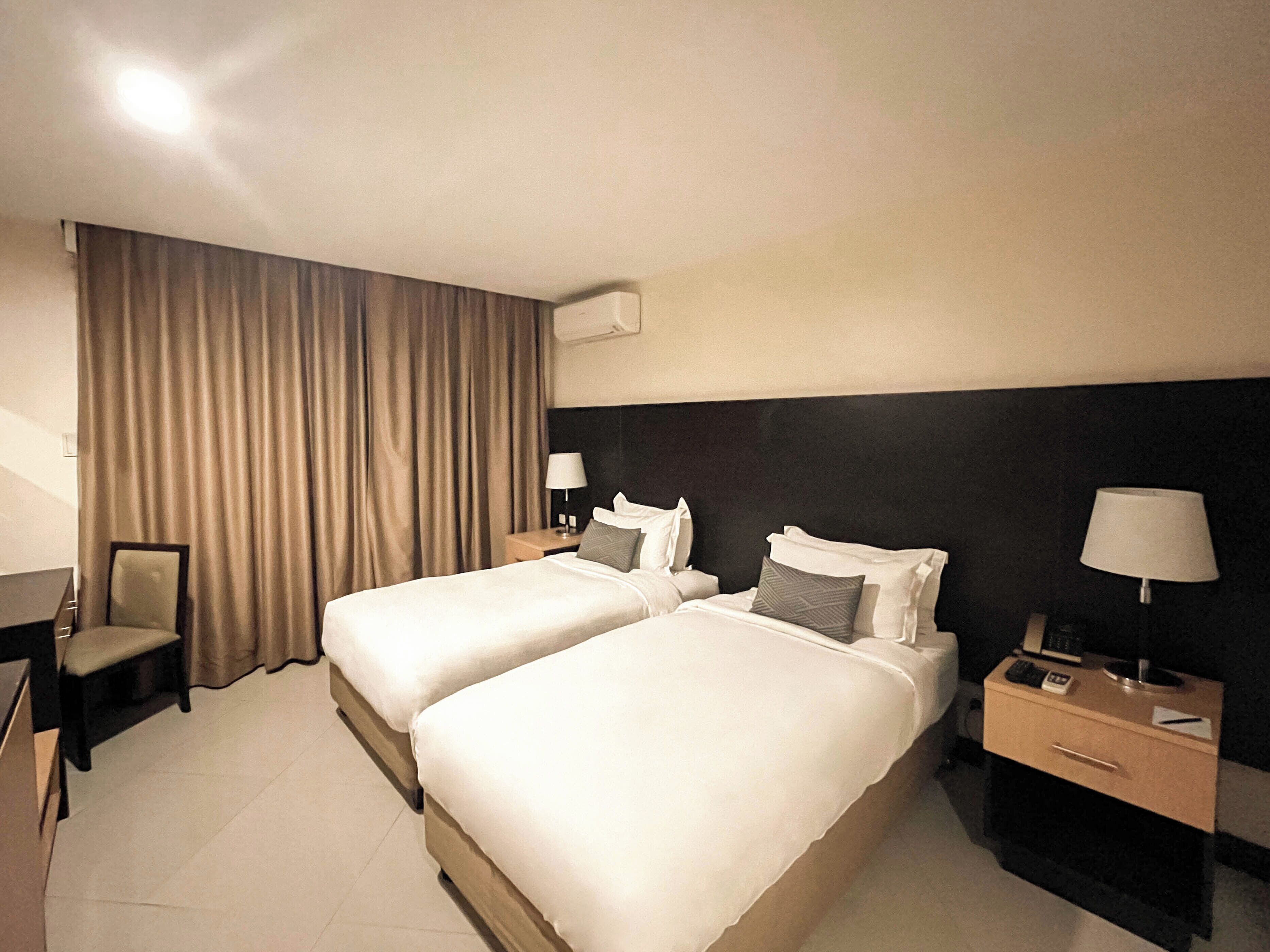 A hotel room with two beds, a desk, a chair, and a nightstand with a lamp.