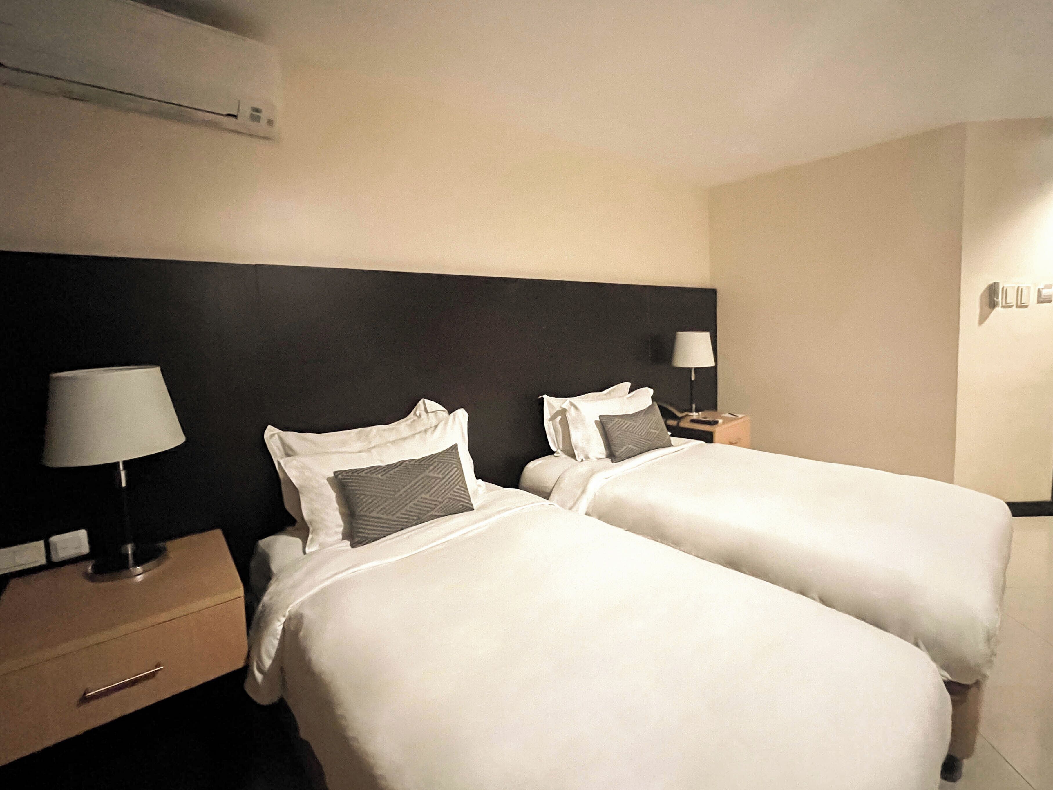 A hotel room with two beds, each with white bedding and pillows, a bedside table with a lamp, and a wall-mounted air conditioner.