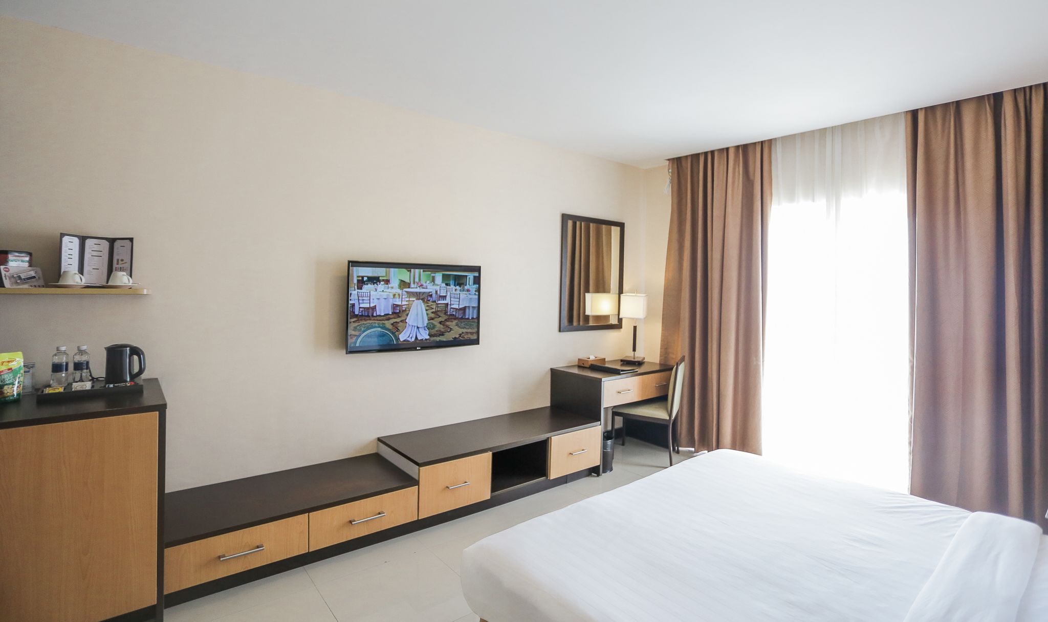 Een hotelkamer met een bed, een bureau met stoel, een aan de muur bevestigde televisie, een lamp en een raam met gordijnen.