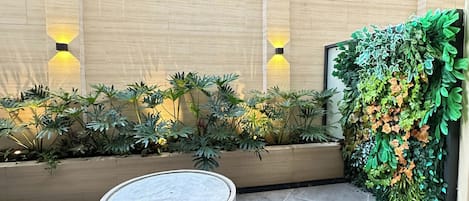Un patio con una jardinera redonda de piedra, suelo de baldosas y una pared con iluminación vertical y vegetación.