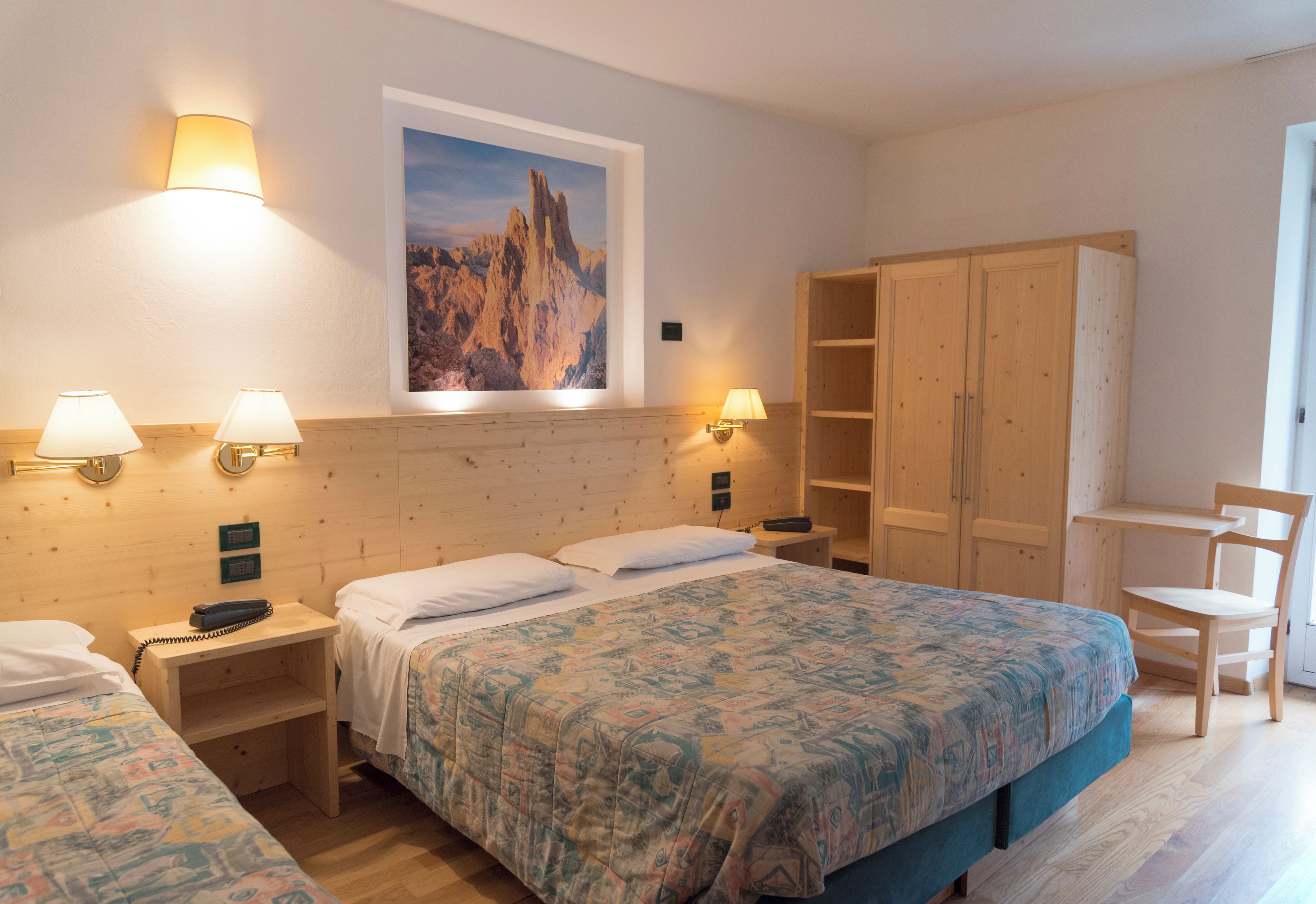 A hotel room with a large bed, two bedside tables with lamps, a wooden wardrobe, a desk with a chair, and a framed picture on the wall.