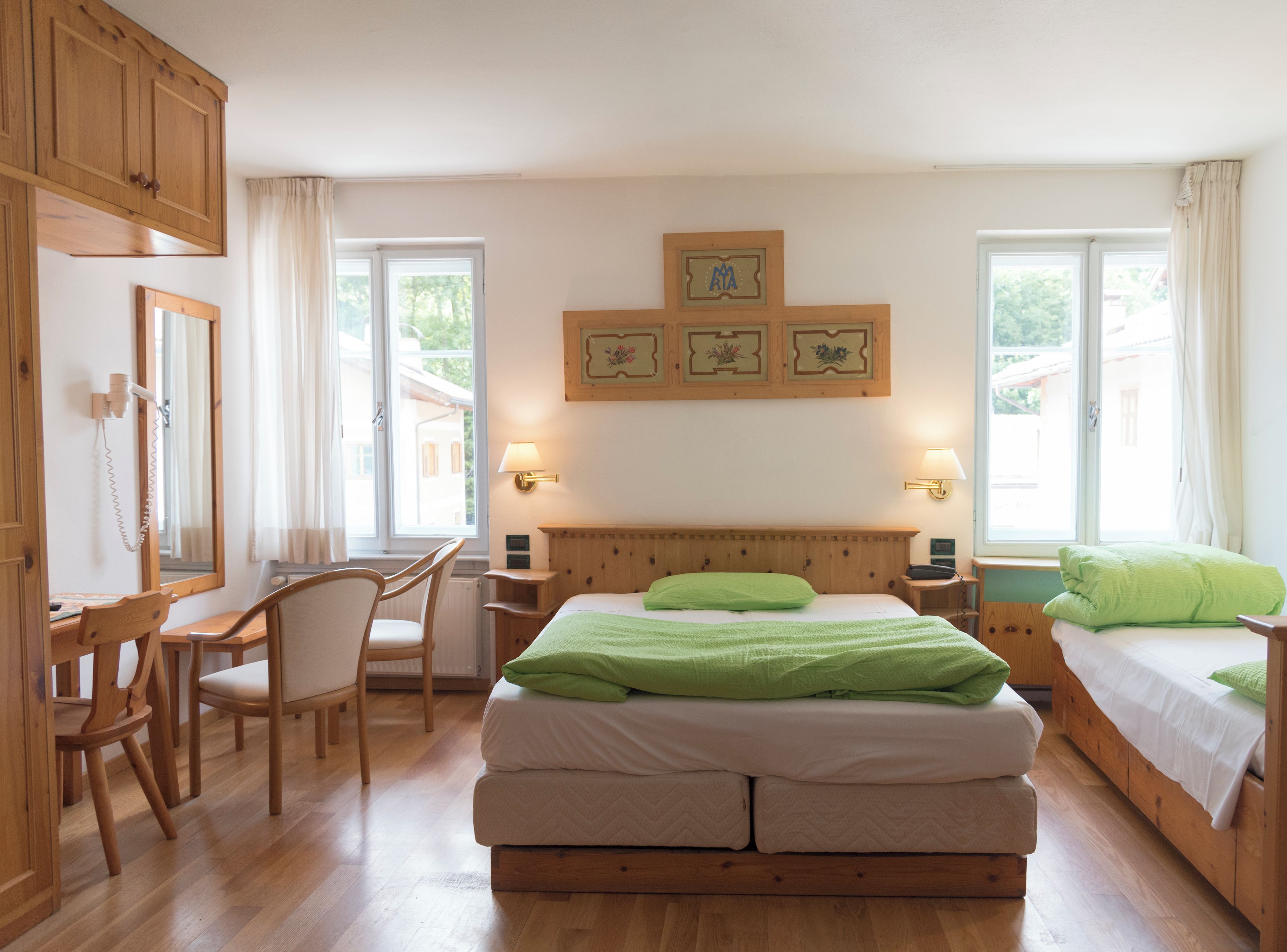 A hotel room with a wooden bed, two bedside tables with lamps, a desk with a chair, and a large window with curtains.
