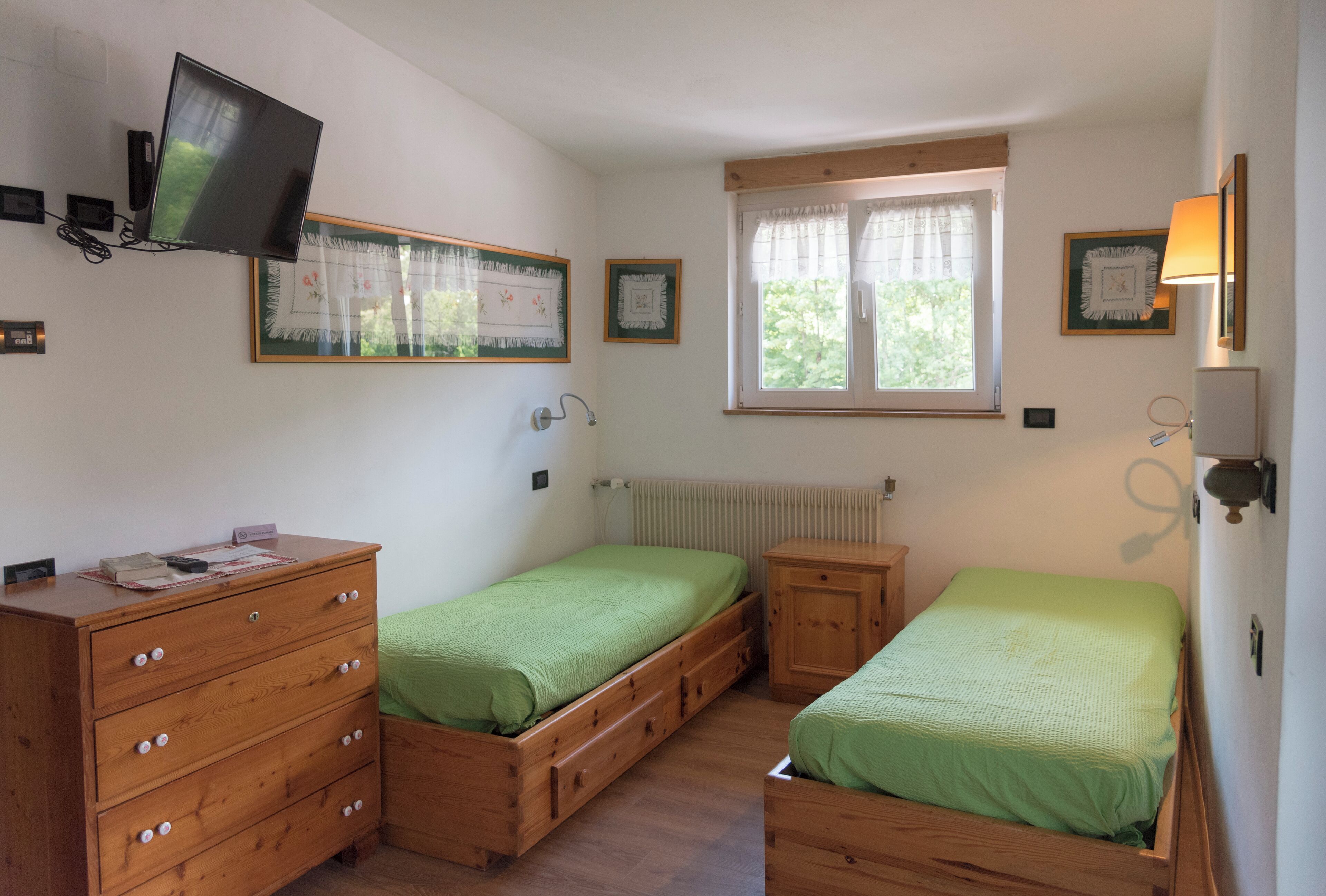 A bedroom with two single beds, a wooden dresser, a television mounted on the wall, and a window with curtains.