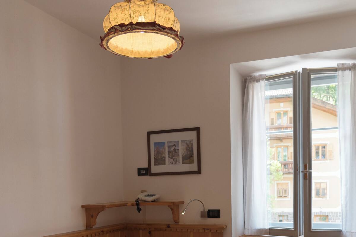 Ein kleiner, gemütlicher Raum mit einem hölzernen Bett, einem Tisch, einem Stuhl, einer Pendelleuchte, einem Fenster mit Vorhängen und einem gerahmten Bild an der Wand.