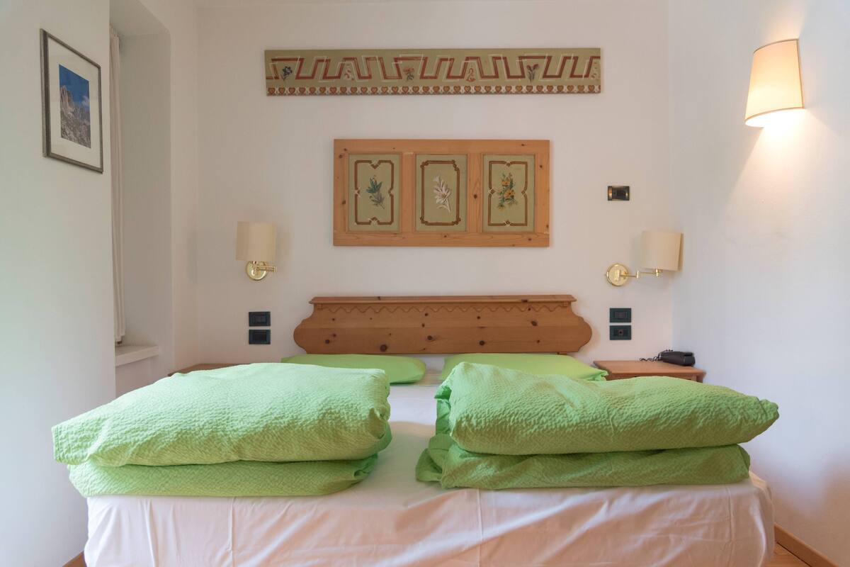 Ein ordentlich bezogenes Bett mit grünen Kissen und einem hölzernen Kopfteil. An der Wand befinden sich Wandleuchten und ein gerahmtes Bild über dem Bett.