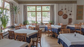 Un comedor con mesas y sillas de madera, ventanas amplias con cortinas, plantas en macetas y utensilios de cocina colgados en la pared.