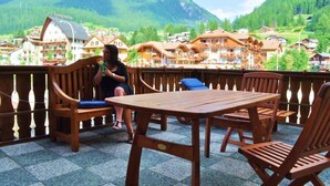 Un espacio de estar al aire libre con madera, que incluye una mesa y sillas, con vistas a un pueblo montañoso con edificios tradicionales.