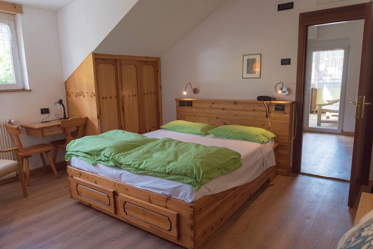 Ein Schlafzimmer mit einem hölzernen Bett, einem Schreibtisch mit Stuhl, einem Heizkörper und einer Tür zu einem Außenbereich.