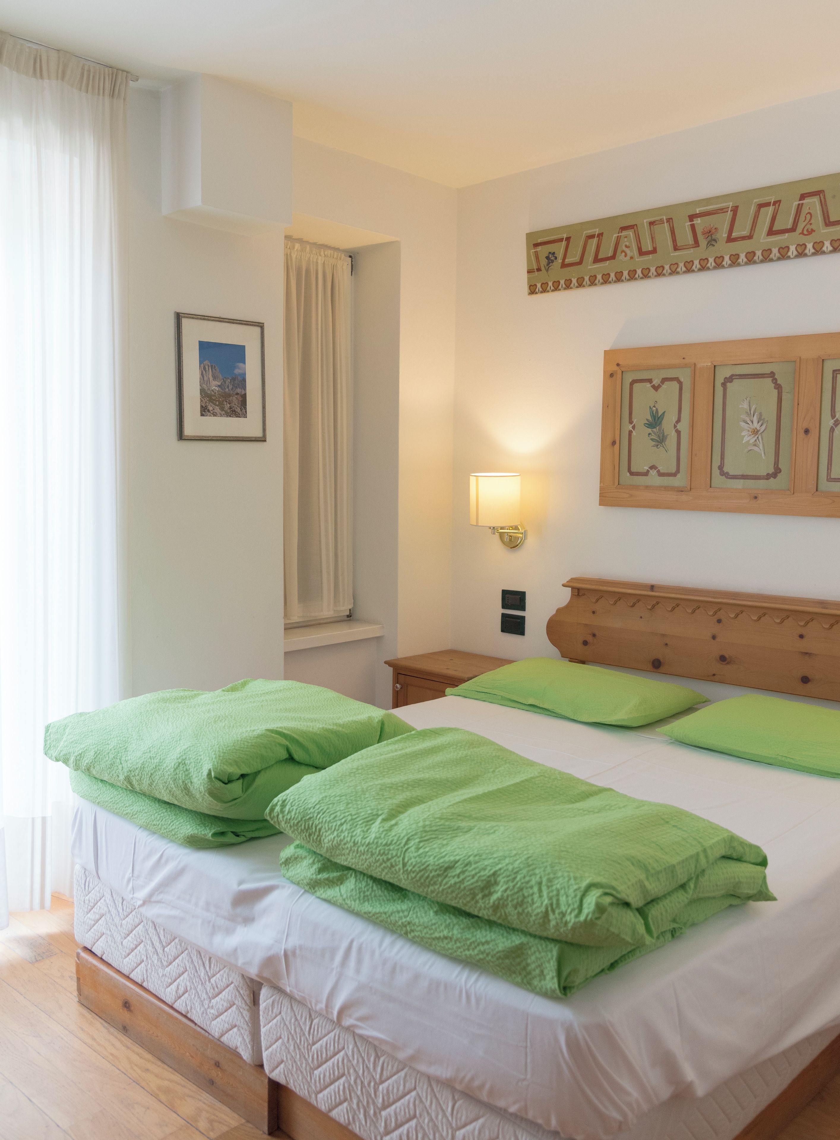 Una cama doble con almohadas verdes y un cabecero de madera, una mesita de noche con una lámpara, un cuadro enmarcado en la pared y una ventana con cortinas.