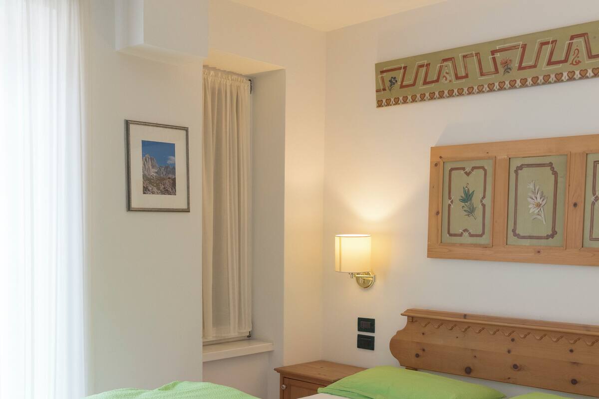 Ein Doppelbett mit grünen Kissen und einem Holz-Kopfteil, ein Nachttisch mit Lampe, ein gerahmtes Bild an der Wand und ein Fenster mit Vorhängen.