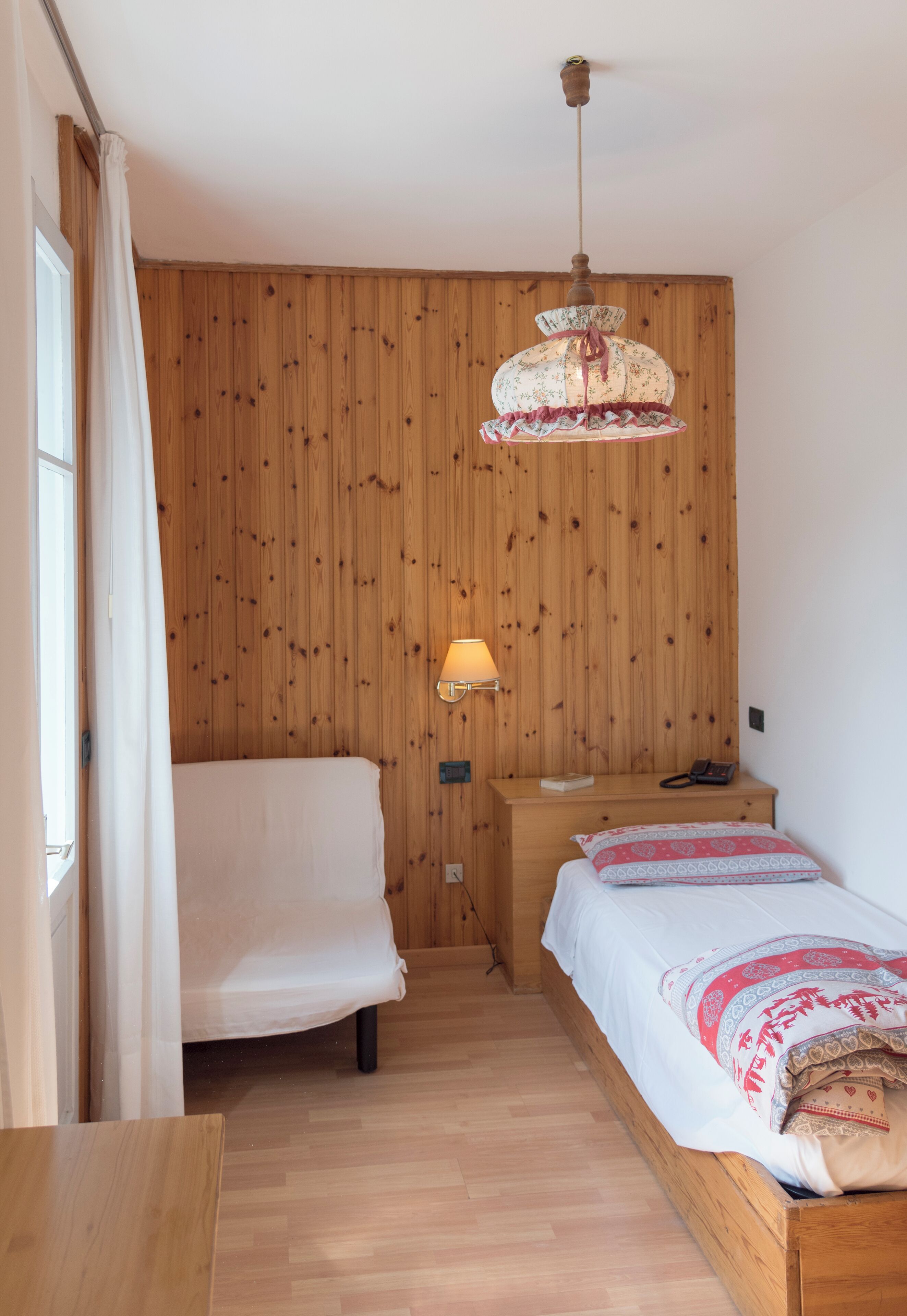 A bedroom with wooden panel walls, a bed with white bedding, a bedside table, a lamp, and a hanging light fixture.