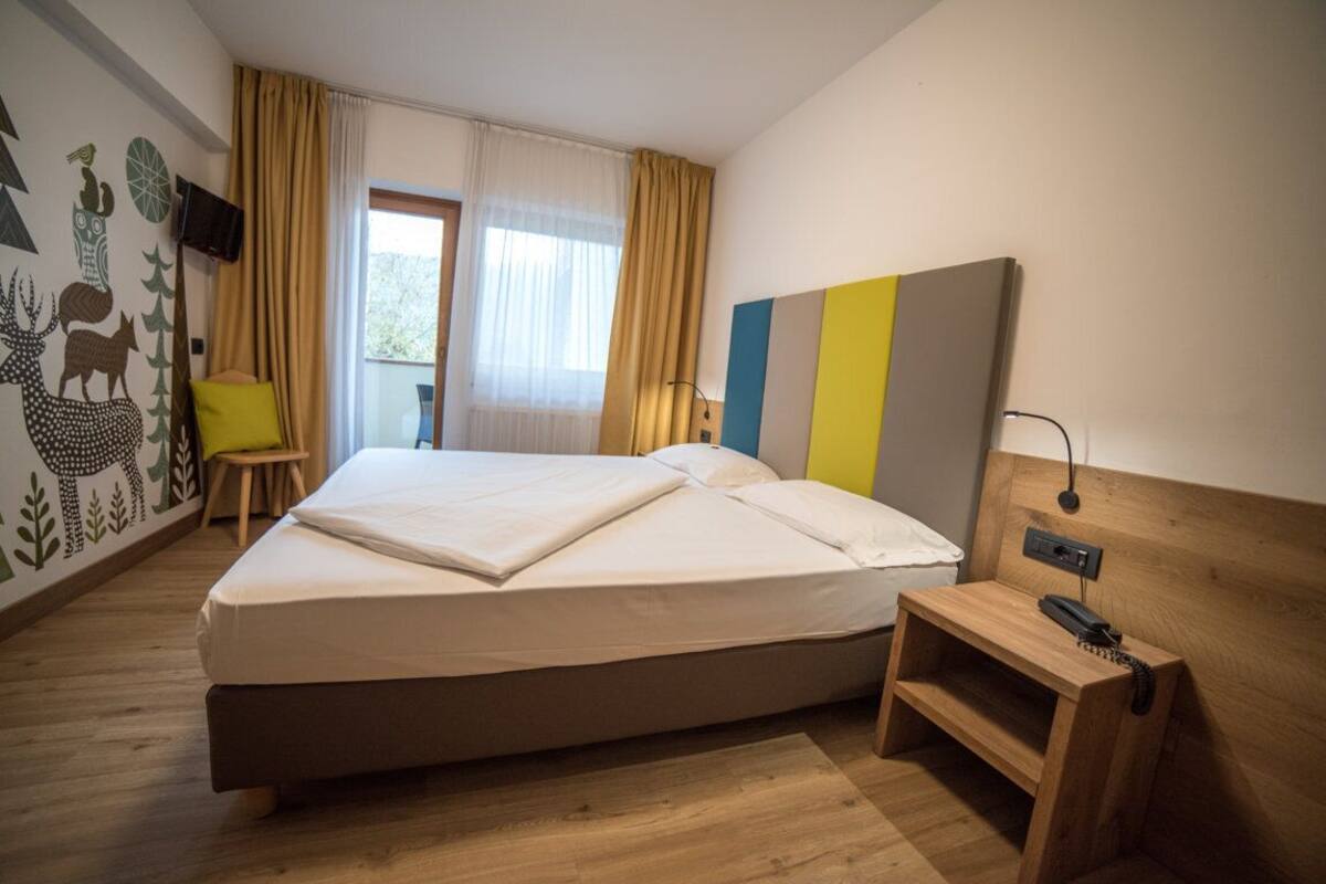 A hotel room with a bed, a bedside table with a phone, a chair, a window with curtains, and a wall with a nature-themed mural.