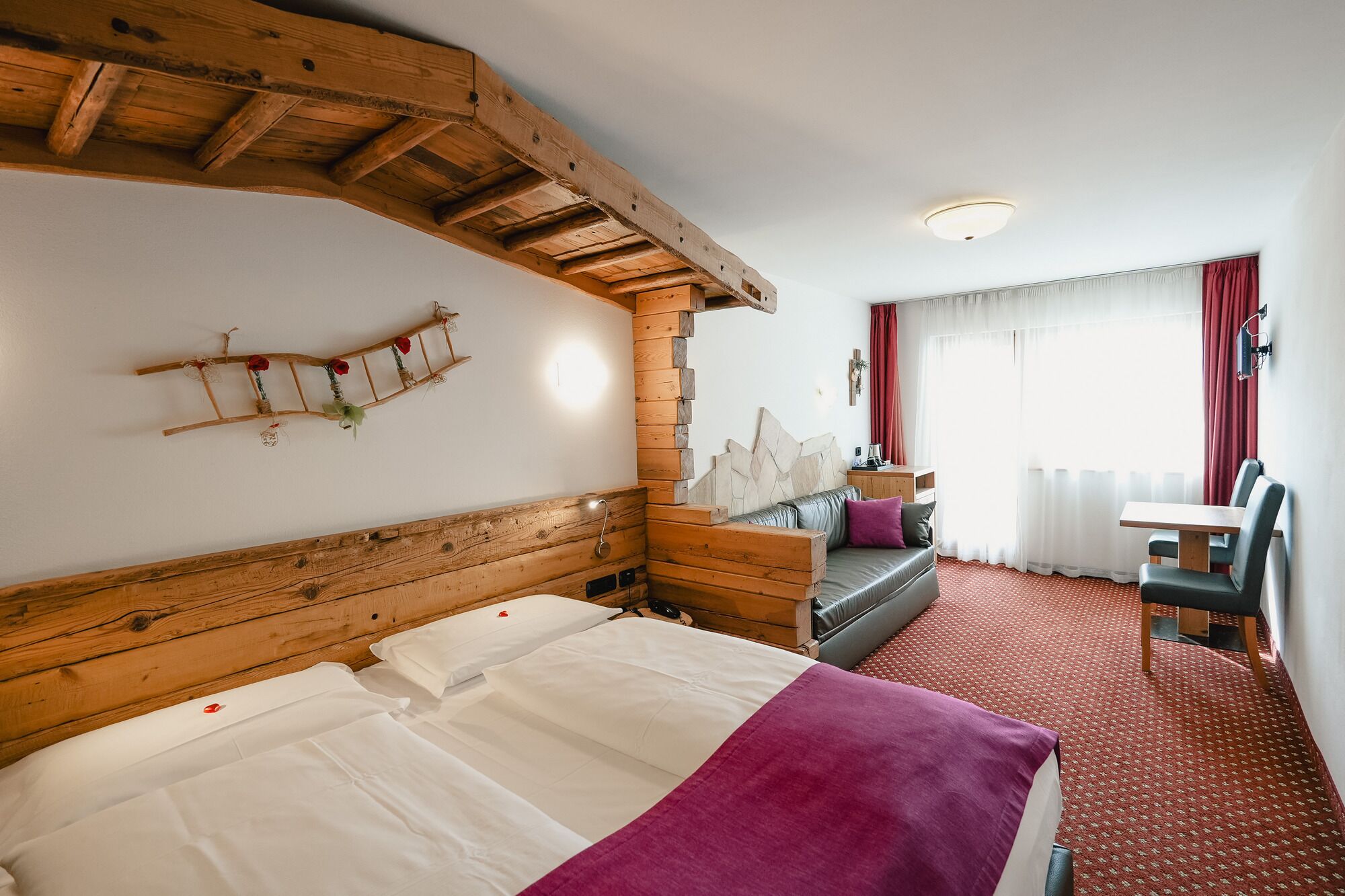 Habitación de hotel con cabecera de madera, cama con ropa de cama blanca y púrpura, una banca de madera, una mesita y una ventana con cortinas rojas.