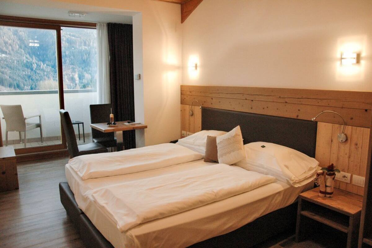 A double bed with a wooden headboard, a bedside table with a vase, and a view of a balcony with chairs and a table.