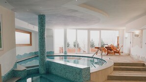 Una amplia piscina interior con suelo de baldosas, un área de descanso de madera con una silla y una mesa, y vistas al exterior a través de amplias ventanas.