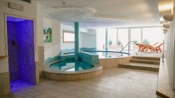 A modern indoor pool area with a central water feature, a wooden deck with lounge chairs, and large windows providing natural light.