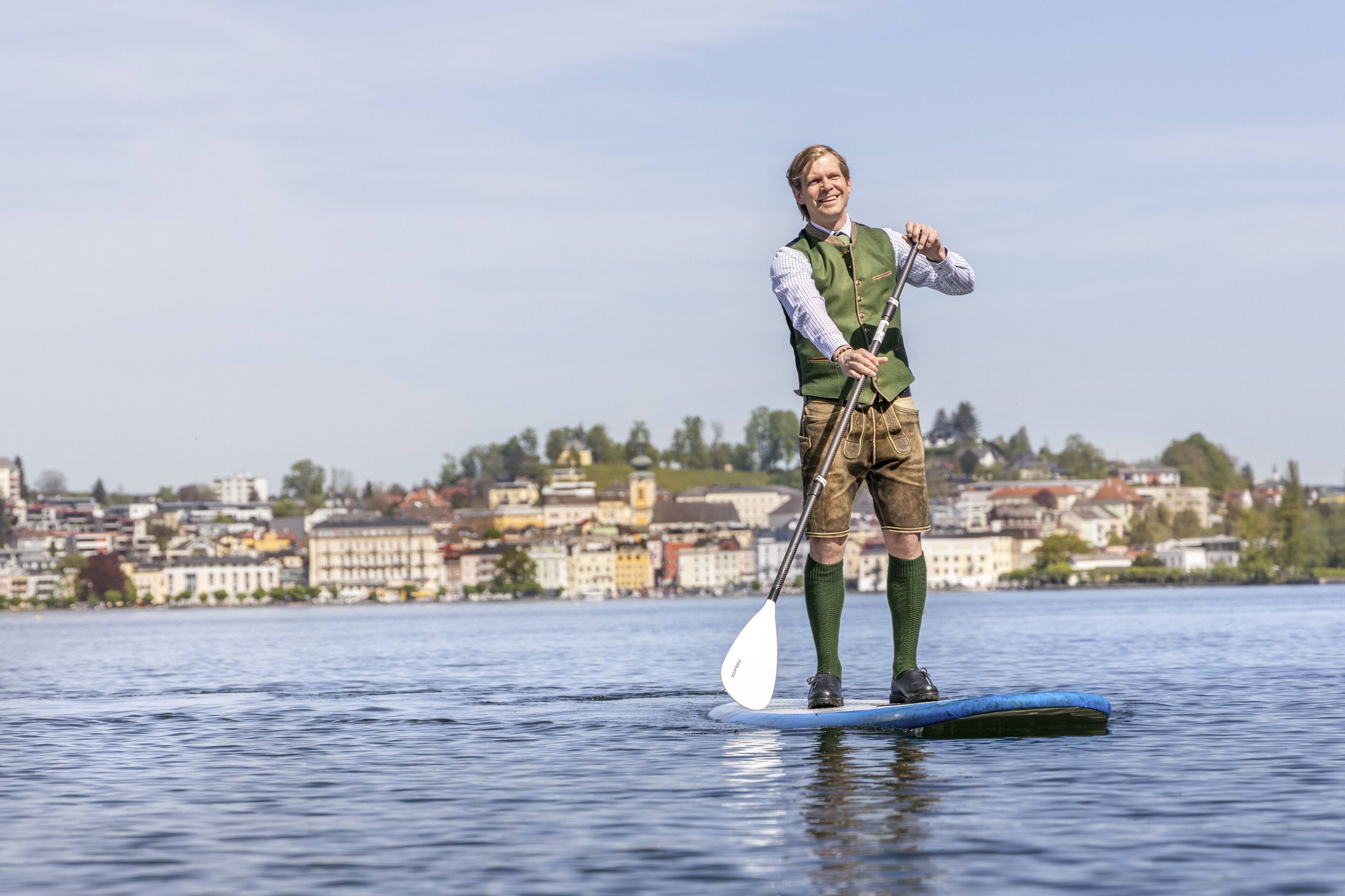 Una persona de pie en una tabla de remo sobre el agua, con un paisaje urbano al fondo.