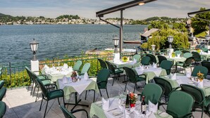 An outdoor dining area with tables and chairs, overlooking a body of water with a distant town. - Landhotel Gasthof Grunberg (Gmunden)