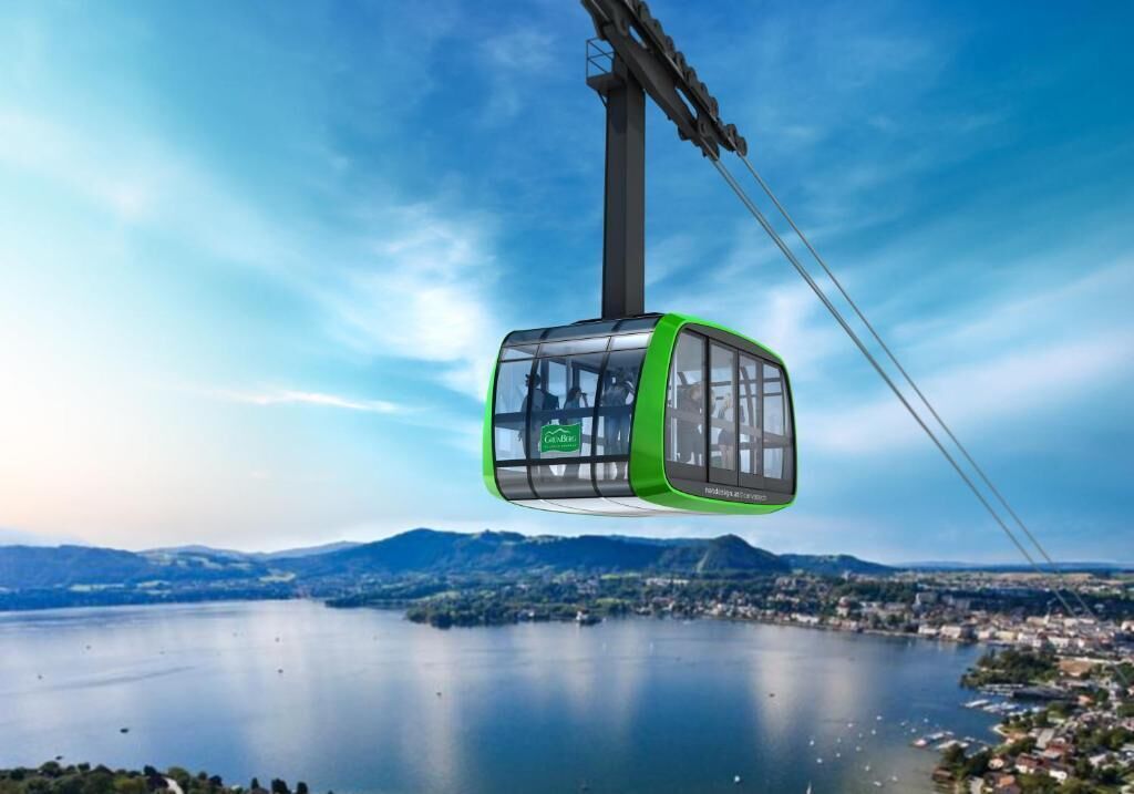 A cable car with a green exterior and transparent windows, suspended by cables against a backdrop of a lake and distant mountains.