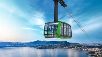 A cable car with a green exterior and transparent windows, suspended by cables against a backdrop of a lake and distant mountains.