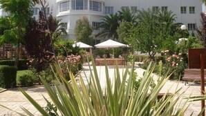 A modern building with a curved facade, surrounded by a landscaped garden with palm trees and flowering plants.