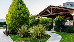 A gazebo with a wooden roof and a stone pathway leading to it, surrounded by well-maintained greenery and a building in the background.