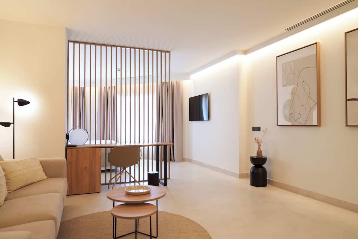 A modern hotel room with a sofa, a dining table, a TV mounted on the wall, and a decorative vase.